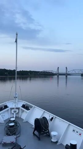 A video shows a view from the bow of a white vessel moving on calm water towards a bridge. The sky is a pale blue with scattered clouds, indicating either dawn or dusk. The bridge, which appears to be a vertical lift bridge, spans a wide body of water. The vessel has several nautical features visible, including a tall pole with an antenna, a winch with coiled rope, and various cleats and capstans. The water reflects the sky and the bridge, creating a serene atmosphere. Trees and a hint of structures are visible on the far bank of the river. The video is shot from a slightly elevated perspective, looking forward and slightly to the right.
