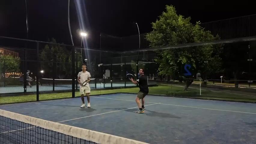Two men are actively engaged in a game of padel on a blue court at night. The man on the right, wearing a dark short-sleeved shirt and shorts, is in the process of serving, having tossed the padel ball into the air with his racket poised for impact. The man on the left, dressed in a white short-sleeved shirt and shorts, stands ready to receive the serve, holding his padel racket.

The padel court is illuminated by bright floodlights, casting light on the players and the playing surface. The court features transparent glass walls at the back and sides, typical of padel facilities, with wire fencing above the glass. A net spans the middle of the court. In the background, other padel courts are visible, also lit, with additional players on them. Large trees with lush green foliage frame the outdoor setting, their outlines stark against the dark night sky. A prominent white "S" is visible on a grassy area behind the court, possibly part of a club logo or sign. The scene captures a moment of active play under artificial light, suggesting a recreational or competitive match taking place in Heroica Puebla de Zaragoza, Mexico.