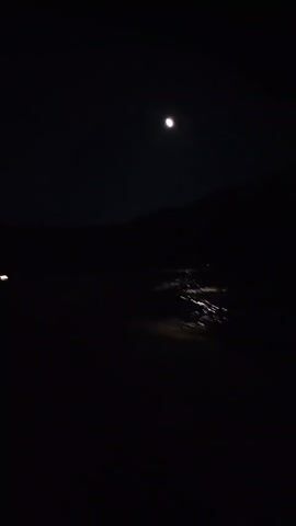 This is a night scene captured in Chuncheon-si, South Korea. The primary subject is the night sky, dominated by a bright full moon. Below the moon, a dark silhouette of mountains or hills is visible. In the mid-ground, a body of water, possibly a river or lake, reflects the light from a distant settlement or a line of streetlights, creating a shimmering effect on the water's surface. There are also a few indistinct lights visible on what appears to be a road or path in the foreground, possibly from vehicles or distant structures. The overall mood is serene and peaceful, with the moonlight casting an ethereal glow over the landscape. The time of day is clearly night, and the weather appears clear, allowing for a good view of the moon and stars. There is no visible text in the media.