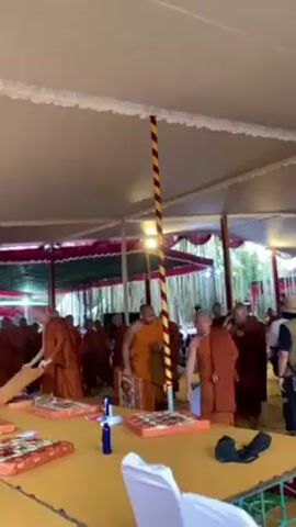 Here is a concise description of the video:

The video shows a gathering of Buddhist monks in a tent. The monks, all wearing orange robes, are preparing for a ceremony or event. They are arranging cushions and small bottles on tables that are covered with a yellow cloth. The tent has a white roof with decorative trim and is supported by poles with yellow and red stripes. Other people can be seen in the background.