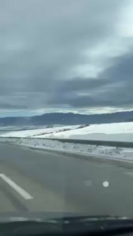 The video shows a view from inside a moving vehicle, likely a car, traveling on a paved road. The road has white lane markings. On either side of the road is a landscape covered in snow. In the distance, there are mountains visible under a cloudy sky. Streetlights are intermittently visible along the roadside. Another vehicle appears briefly ahead on the road. The interior of the car is partially visible at the bottom of the frame, including the dashboard.