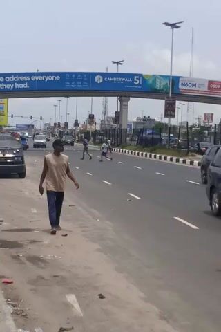 The vertical video captures a man walking along the dusty, unpaved shoulder of a multi-lane highway in Eputu Town, Nigeria, during bright daylight.

The man, appearing in the foreground, is wearing a light-brown short-sleeved t-shirt, dark blue trousers, a dark baseball cap, and light-colored shoes. He walks with a steady pace, looking forward and slightly to his right. His expression appears neutral. Scattered litter and debris are visible on the ground next to him.

The asphalt road, marked with white lane lines, stretches into the distance, occupied by several vehicles, including a dark sedan closest to the camera and other cars further back, suggesting moderate to heavy traffic flow.

A large blue and white billboard structure spans across the highway overhead. Visible text on the billboard includes the slogan "OWN THE ADDRESS EVERYONE WISHES THEY HAD.", "CAMBERWALL 5", and "N60M+" at the top right. Further down the road, another individual can be seen walking away from the camera on the roadside. The sky is clear, indicating sunny weather.