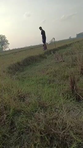 Two individuals are in a grassy field with a channel of water or a ditch in the foreground. The person in the foreground, a male, is walking away from the camera. He is wearing a dark t-shirt and shorts. In the background, a second person, likely a child, is on the far side of the channel. This person is wearing a blue and white striped shirt and dark shorts and is pulling a white object, possibly a kite or a toy, behind them. The environment appears to be rural, with trees visible in the distance and a bright, hazy sky. The lighting suggests it is either early morning or late afternoon.