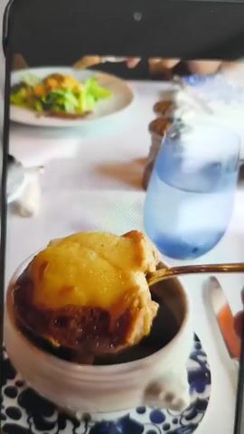 The video shows a close-up of a bowl of French onion soup being stirred with a gold spoon. The soup is in a white bowl with a blue and white patterned saucer. The top of the soup has a melted cheese topping and a piece of bread. Next to the bowl is a light blue glass of water. In the background, there is a salad on a white plate and the hands of a person. The table is covered with a white tablecloth. The person stirring the soup is not fully visible.