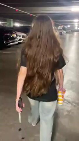 The video shows a person with long brown hair walking through a parking garage. The person is wearing a black t-shirt, light gray pants, and black shoes, and carrying a McDonald's cup. They appear to run in the direction of a black car, turn around to face the camera, and fall. The parking garage is well-lit and lined with cars. The person seems to be slightly disoriented.