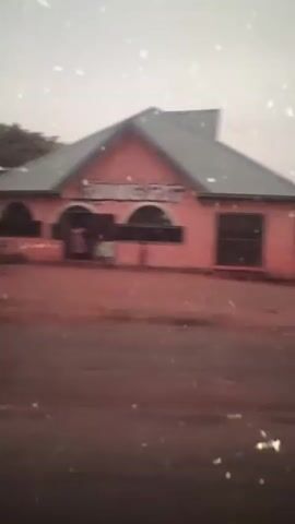 The video shows a view from the inside of a vehicle moving down a red dirt road. Outside the window is a pink building labeled "Running Spot". The road is wet, and there are drops of water on the window. There are trees and other buildings visible in the background. The video is shaky and the focus is blurry.