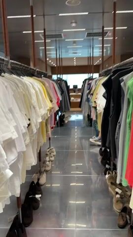 The video shows a clothing store aisle. The camera is positioned at eye level looking down the aisle. On both sides of the aisle are clothing racks filled with various garments, mostly t-shirts and some outerwear. The floor is reflective and appears to be made of metal or polished concrete, reflecting the clothing racks and overhead lighting. In the background, two individuals, a man and a woman, are walking away from the camera and deeper into the store. The woman is wearing a pink baseball cap, a white top, and jeans. The man is wearing a grey jacket and pants. Further in the background, another person, appearing to be a female employee, is visible standing behind a counter or display. The overall impression is a modern, minimalist retail environment.