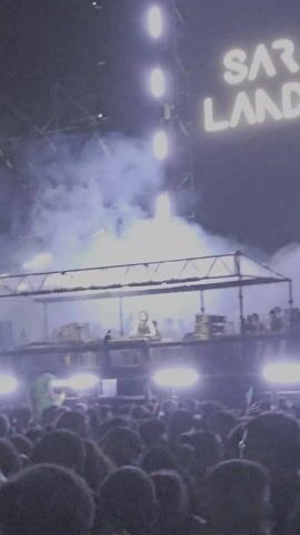 The video shows a concert or music festival featuring the artist "SARA LANDRY" as displayed on a large sign behind the stage. The lighting is intense, with spotlights and smoke enhancing the atmosphere. A DJ or performer is visible on a raised platform, surrounded by equipment. Below, a large crowd of people faces the stage, indicating an active and engaging performance.