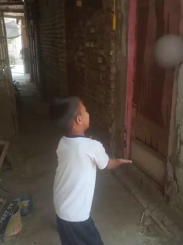 A boy in a white t-shirt and black shorts is playing with a ball. He throws the ball up in the air and catches it, then throws it up again and catches it. He is in a narrow alleyway with brick walls and a dirt floor. There is a wooden door in the background. The video is recorded from a low angle.