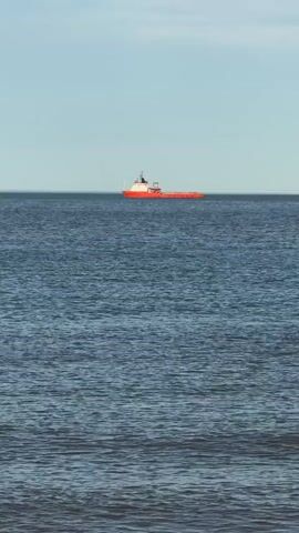 A bright orange and white ship is visible on the horizon across a vast expanse of blue water under a clear sky. The ship appears to be a supply vessel or a similar type of workboat, characterized by its long, flat deck. The water exhibits gentle ripples, suggesting a calm sea. The perspective is from a low vantage point, possibly from a beach or a low-lying coastline, as indicated by the very bottom of the frame showing a dark, slightly textured area that could be sand or pebbles. The overall scene is serene and expansive, emphasizing the isolation of the vessel in the open sea.