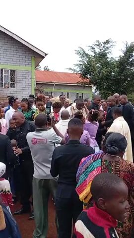 Here is a description of the video:

The video shows a large group of people gathered outside a building. They appear to be celebrating an event, as someone is being carried in a chair above the crowd. Many people are taking pictures and videos. There are a variety of clothing styles visible in the crowd, including traditional African clothing, suits, and casual wear. The buildings in the background appear to be simple, possibly a school or community center. The weather appears to be overcast.