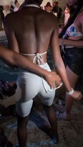 The video shows a person with dark skin in white shorts and a matching top dancing by a pool. Another person places their hand on the dancer's buttocks while they dance. Other people are in the background near the pool, some dancing. The scene appears to be a party or social gathering near a pool at night.