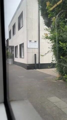 The video shows a view from a window of a moving vehicle, looking out at a street scene. On the left is a light-colored building with dark trim and several windows. A sidewalk with paving stones runs in front of the building. To the right of the sidewalk is a dense green hedge. A street sign with a blue rectangle and a white line is visible, followed by a circular sign with two figures, one adult and one child. Another sign with a red circle and a horizontal line is also visible. Trees and more greenery line the street. The sky appears overcast.