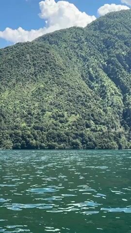 A white motorboat with two people aboard speeds across a dark green lake, creating white wakes. The boat is moving from left to right in the frame. Behind the lake, a large, densely forested green mountain rises steeply under a bright blue sky dotted with white clouds. The mountain is covered in trees and has a rugged texture. A narrow, winding road is visible partway up the mountainside. The sun is shining, and the water is choppy.