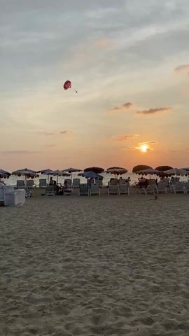 The video shows a beach scene at sunset. In the foreground, there is a sandy beach with rows of beach chairs and umbrellas. Some people are sitting on the chairs, seemingly enjoying the view. In the background, the sun is setting over the ocean, creating a warm, orange glow. A person is parasailing in the sky. The sky is filled with clouds.