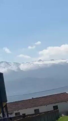 The video shows a rural scene with snowy mountains in the background, under a blue sky with scattered clouds. In the foreground, there's a corrugated metal roof, partially obscured by a gray wall with graffiti. A large, cylindrical metal tank is also visible above the roof. The camera pans slightly, revealing a bare tree against the backdrop of the snow-covered mountains. The overall atmosphere seems windy.