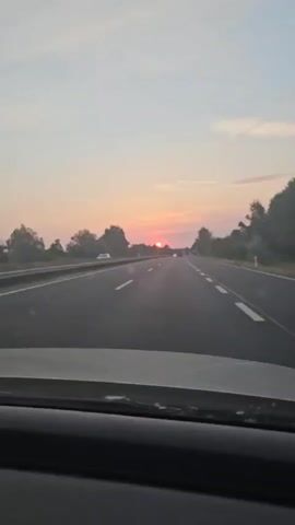 A first-person perspective video captures a sunset view from inside a moving vehicle. The road ahead is paved and marked with white lines, indicating a highway or major road. Trees line both sides of the road, and the sky is a soft gradient of pink and orange, with the sun visible near the horizon. The dashboard of the vehicle is partially visible in the foreground. There are other vehicles on the road in the distance, and a few signs are visible along the side of the road. The overall scene depicts a peaceful journey during sunset.