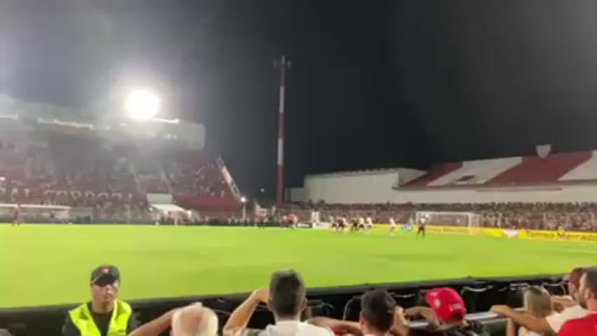 The video shows a soccer game at night from the perspective of a spectator in the stands. The stadium is full of fans, mostly wearing red and white. A tall red and white striped tower stands near the side of the field. Players in white uniforms are lined up near the goal. The field is brightly lit. Security personnel wearing yellow vests are visible in the foreground. Other spectators can be seen in the foreground, watching the game intently. Some have their hands on their heads.