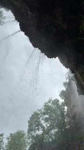 The video shows a view looking up at a waterfall from underneath its overhang. Water streams down from the dark rock face, creating a misty effect in the air.  Trees with green foliage are visible at the bottom of the frame, partially obscured by the waterfall's spray. The sky is overcast and gray. The angle is slightly tilted upward.