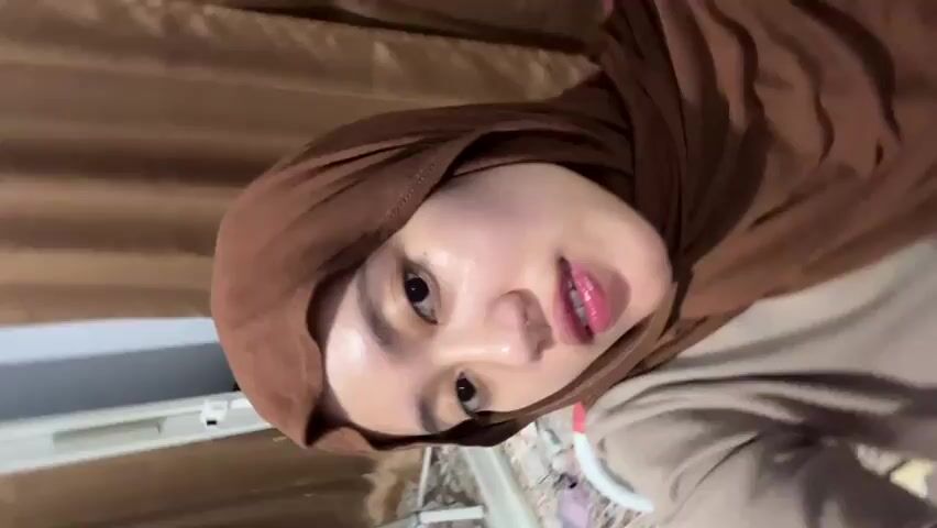 A woman wearing a brown hijab and a light-colored top looks directly into the camera. She has glossy pink lips. She then smiles, revealing silver braces on her upper teeth. Her face appears shiny, possibly due to moisturized skin. A brown curtain is visible in the background.