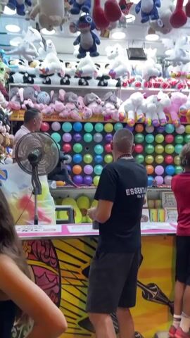 At a carnival game booth, a man in a light pink shirt stands behind a counter on the left, holding a white plush toy. In front of the counter, a man in a black t-shirt with "ESSENTIALS" printed on the back faces away from the camera, holding a dart. To his right, a young person in a white t-shirt looks at the game. Behind the counter, a wall is covered with rows of colorful balloons. Above the balloons, shelves are filled with various plush toys, including unicorns, pandas, elephants, and a blue Stitch character. A black fan sits on the counter in front of the man in the pink shirt. In the foreground, the shoulder of a person in a pink shirt is visible. The environment appears to be an indoor carnival or amusement park setting.