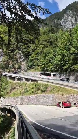 A video shows a winding road with a bus and several cars driving along it. The road is carved into the side of a mountain, with lush green trees covering the slopes. In the foreground, there are two people standing next to a red car that is parked on the side of the road. The sky is blue with some clouds. The video appears to be taken from a higher vantage point, looking down on the road and the surrounding landscape.