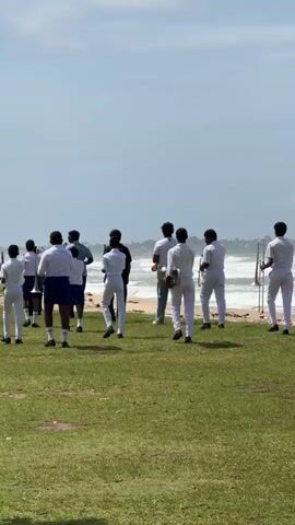 A group of young people in white uniforms and some in green uniforms are walking on a grassy field near the ocean. Some of them are carrying musical instruments, possibly for a marching band. The sky is cloudy, and the ocean has waves. The overall scene appears to be an outdoor practice or rehearsal.