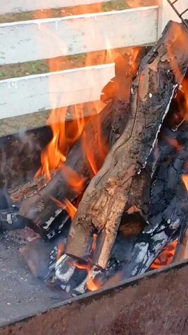The video shows a close-up view of burning logs in a fire pit. The logs are a mix of dark, charred wood and brighter, orange flames. Smoke is also visible, rising from the fire. In the background, a white railing or fence can be seen. The overall impression is of a warm, outdoor setting with a campfire.