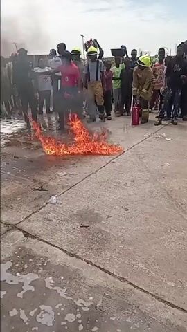 This media file captures an outdoor fire safety demonstration or training exercise in Katabu, Nigeria, under bright daylight conditions. A small, controlled fire burns actively on a concrete surface, emitting thick, black smoke.

A man wearing a red polo shirt, dark trousers, and a black cap, with an ID badge clipped to his shirt, is the central figure attempting to extinguish the flames. He initially runs towards the fire and then proceeds to pour water onto it from a small plastic bottle. Despite the water, the fire continues to burn, indicating that it is likely a liquid fuel fire, for which water is not an effective extinguishing agent.

A group of people, including several individuals in firefighter suits with bright yellow helmets, watch the demonstration. One firefighter, in a khaki/tan suit with "AG FIRE" clearly visible on the back, walks towards the fire while carrying a red fire extinguisher. Other observers include people wearing white shirts with red cross symbols (suggesting Red Cross personnel) and uniformed individuals who appear to be police or security, along with numerous other civilians and children.

The concrete ground around the fire is wet, possibly from previous attempts or the poured water, and some foamy residue is present. The overall atmosphere appears calm and instructive, typical of a controlled public safety event rather than an emergency.