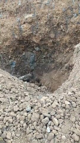 The video shows an excavator digging a large hole in a hillside. The hole is surrounded by piles of dirt and rocks. There are trees and vegetation on the hillside above the hole. The excavator is yellow and appears to be in good condition. There is a person standing near the excavator, but their face is not visible. The video is shot from a distance, so it is difficult to make out fine details. The overall impression is one of construction or excavation work.
