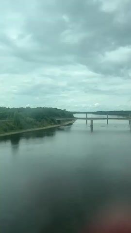 A video shows a wide river with a bridge spanning across it. The sky is overcast with heavy clouds. The riverbanks are lined with trees and vegetation. There is a small boat on the water to the right.