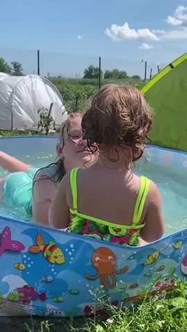 This media file captures two young girls playing in a small, inflatable swimming pool on a sunny day in Pesnevo, Russia. The pool is decorated with colorful marine life, including fish and an octopus. One girl, with blonde hair and wearing a teal swimsuit, is partially submerged in the water, smiling and looking towards the other girl. The second girl, with brown curly hair and wearing a neon green and floral swimsuit, has her back to the camera, facing the blonde girl. Both appear to be enjoying themselves in the water.

The scene is set outdoors, with lush green grass surrounding the pool. In the background, a white, tent-like structure or greenhouse is visible, along with a green tent or tarp. Beyond that, there are trees and a hint of a fence, suggesting a backyard or garden setting. The sky is a clear, bright blue, indicating pleasant weather, and the sun is high, suggesting it's daytime. The overall mood is one of playful enjoyment and summer fun. No visible text is discernible in the media file.