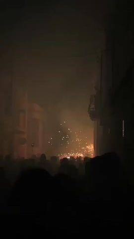 The video shows a crowd of people in a dark, narrow street. There is smoke and a bright display of fireworks or sparks in the distance. People are silhouetted against the light, with some looking towards the event. The buildings on either side of the street are dimly lit.
