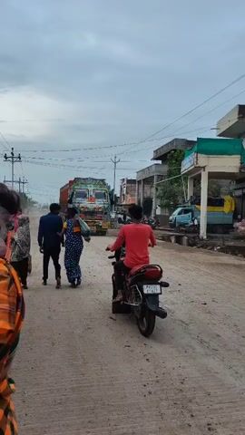 The video shows a busy street in what appears to be a rural or semi-urban area. A large, brightly decorated truck is driving down a dirt road. Several people are walking on the side of the road, and two individuals are riding motorcycles. Buildings are visible on the right side of the street, and power lines crisscross the sky. The weather appears to be overcast. The truck is the main focus of the video, with its colorful decorations suggesting it is a commercial vehicle, possibly from India. The scene depicts everyday life on a local road.