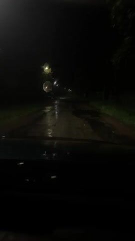 The video shows a first-person view from inside a vehicle driving down a dark, wet road at night. Streetlights illuminate patches of the road, revealing puddles and reflections. Trees and grass are visible along the sides of the road, and in the distance, houses and parked vehicles can be seen. The windshield of the vehicle is wet, with water droplets visible. The overall impression is of driving through a rural or suburban area during a rain shower.