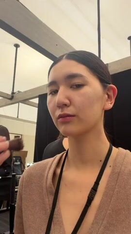 A makeup artist applies powder with a brush to a young woman's face. The woman has her hair pulled back, is wearing a brown sweater with a black strap around her neck, and is looking directly at the camera. The makeup artist is partially visible, wearing a black shirt. The scene appears to be backstage, with other individuals and makeup stations visible in the background. The area is lit by overhead fluorescent lights.