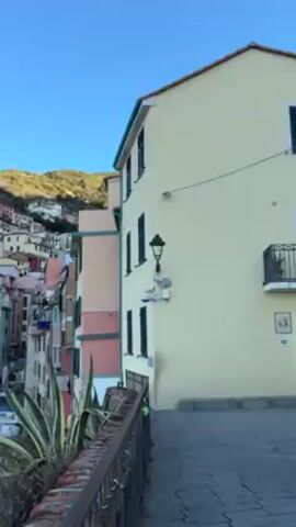 The camera pans across the colorful town of Riomaggiore, Italy. The buildings are stacked up the side of a mountain with the clear blue sky in the background. The buildings are different colors, including red, pink, yellow, and white. Small boats are docked along the harbor, and people can be seen walking near the boats. The camera slowly pans up to capture the full scope of the hillside town.