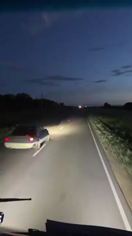 The video shows a view from inside a vehicle traveling on a two-lane highway at dusk. The sky is a deep blue with a few clouds, and the road ahead is lit by the vehicle's headlights and the headlights of other cars. In the distance, a car is visible with its taillights illuminated, and further ahead, there appear to be more vehicles. The road is bordered by dark, silhouetted trees on the left and a field of tall grass on the right. The white lines marking the lanes are clearly visible under the headlights. The perspective is from the driver's or passenger's seat, with the dashboard and windshield wipers partially visible in the foreground. The overall impression is one of nighttime travel on an open road.