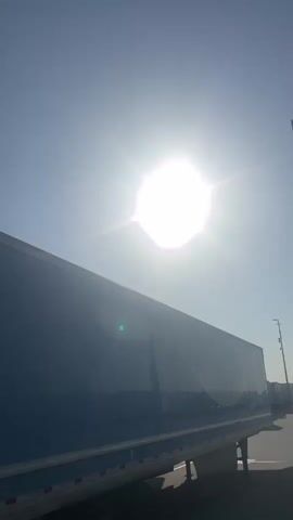 The video pans upward towards a bright sun against a clear blue sky. In the foreground, the top of a dark blue semi-trailer truck is visible, angled upwards. Several streetlights with square fixtures at the top are seen along the side of what appears to be a road. The sun is very bright, causing lens flare and obscuring details in the sky.
