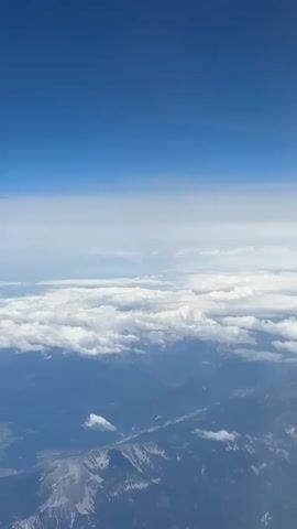 The video shows an aerial view of mountains and clouds. The sky is a clear blue, with a layer of white clouds below. The mountains appear rugged and covered in green vegetation, with some snow-capped peaks visible. The lighting suggests it is daytime, possibly late morning or early afternoon. The perspective is from an aircraft window, looking down at the landscape. There are no people or man-made objects clearly visible.