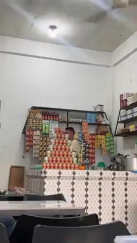 The video shows the interior of a small, simply furnished eatery. The camera slowly zooms out from the counter area towards the dining space. Behind the counter stands a man, presumably the shopkeeper, amidst an array of goods. A pyramid of canned goods is prominently displayed on the counter, and various snacks and drinks hang on the wall behind the counter.  Shelves above the counter hold more merchandise. In the dining area, there are tables and chairs. One person is seated at a table, with another person visible briefly. The ceiling is plain, with a fan. Natural light filters in from the exterior. The overall ambiance appears casual and functional.