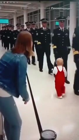 This media file shows a group of people in what appears to be an airport or a similar public indoor space with a polished floor and modern architecture. In the foreground, a woman with reddish-brown hair, wearing a denim jacket and light-colored pants, is leaning down, facing away from the camera. A toddler in a red outfit and white shoes is walking away from her, towards a line of individuals in uniform.

The individuals in uniform are wearing dark jackets with badges and hats, resembling pilots or airline personnel. They are walking in a single file line. As the toddler walks towards them, one of the uniformed individuals bends down to interact with the child.

The lighting suggests it is daytime, but there are no visible windows or cues to indicate the weather outside. The setting appears to be a hallway or concourse, possibly an airport terminal, given the uniforms and the general environment. There is a black retractable belt barrier visible in the foreground.

The overall emotion conveyed is one of tenderness and interaction, with the uniformed personnel showing attention to the child. The toddler's stance and movement suggest a child exploring or walking towards the approaching line of people.

Visible text overlays include "28", "Сообщество", "Подписки", and "Рекомендации", suggesting the media is being viewed on a platform with these interface elements, likely Russian-language based. The specific location of Kamianske, Ukraine, is provided as context but not directly evident from the visual content itself.