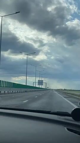 The video shows a view from inside a car driving on a highway. The sky is overcast with dark clouds, and the road appears wet. Street lights line both sides of the highway, and a green guardrail is visible on the left. Other cars are traveling in the same direction. The dashboard of the car is visible at the bottom of the frame.