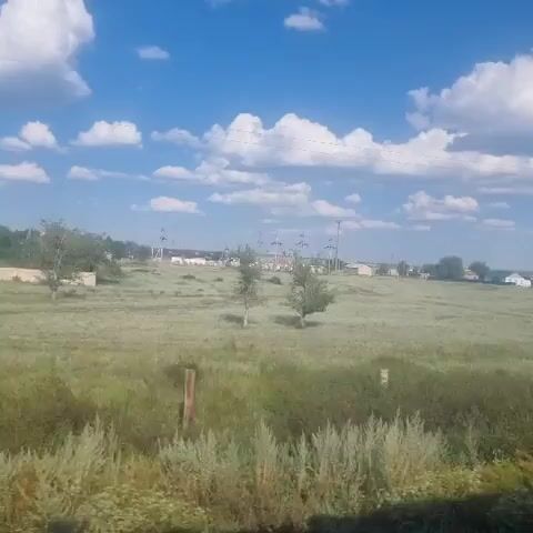 The video shows a landscape with a bright blue sky and scattered white clouds. A grassy field occupies the foreground and middle ground, with some bushes and wooden posts visible. In the distance, there are several houses and what appear to be oil pumps or similar industrial structures. Two tall electricity pylons are prominent features in the landscape, one closer to the foreground on the right, and another further back on the left. A few people, likely children, are visible in the middle ground of the field, near a bucket. A white car is parked in the distance. The overall impression is of a rural or semi-rural setting on a sunny day.