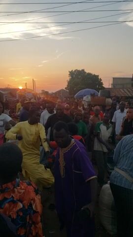 This media file captures an outdoor gathering of people in Kakuri Gwari, Nigeria, during sunset. The warm, orange glow of the setting sun is visible in the background, illuminating the sky and casting a soft light on the scene. A crowd of individuals, dressed in various casual attire including shirts, dresses, and traditional clothing, are congregated. Some are looking towards the camera, while others are engaged in conversation or observing the surroundings. The atmosphere appears to be lively and communal, with a sense of shared experience.

Several objects are discernible within the frame, including plastic crates and what appears to be a tent or canopy, suggesting a market or a community event. A banner with the text "POS" is visible, likely indicating a point of sale or financial service. The subjects exhibit a range of expressions, from smiles to more neutral gazes, contributing to the dynamic feel of the captured moment. The setting appears to be a dusty or unpaved area, typical of many outdoor spaces in Nigerian communities. The overall impression is one of a vibrant local gathering taking place as day transitions into evening.