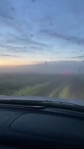 The video shows the view from inside a vehicle driving on a dirt road. The sky is cloudy with a hint of sunrise colors. There is fog or mist obscuring the landscape, which appears to be fields or tall grass. The vehicle's headlights are on and illuminate the road ahead. In the distance, the red taillights of another vehicle are visible. The dashboard of the vehicle is in the foreground, with a sticker on it. The video captures the motion of the vehicle as it drives through the foggy landscape.