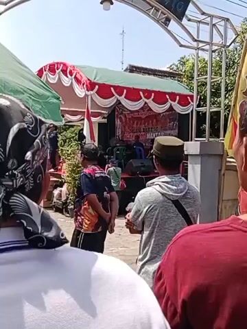 A medium shot shows a group of people gathered around a makeshift outdoor stage. The stage is decorated with red and white fabric. A banner on the stage displays "REPUBLIK INDONESIA" and "JALAN BARENG MANTAR." There are several people on the stage, including musicians playing instruments, and some seated. In front of the stage, several individuals are visible, some looking towards the stage, while others are partially obscured. One person in the foreground wears a red shirt, another a grey hooded sweatshirt with a black strap across their chest, and a third wears a dark shirt with a colorful graphic. A person in a black cap and a blue shirt with a graphic stands near the front. To the left, a motorcycle is parked. The environment appears to be an outdoor gathering with trees and a clear sky visible. The overall scene suggests an event or performance taking place.