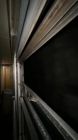 The video shows a view from inside a train car looking out a partially open window. The window frame is metal and appears somewhat weathered. Outside the window, it is completely dark, suggesting it is nighttime or the train is in a tunnel. The interior of the train car is visible on the left side of the frame, including a hallway with wooden doors and some metallic fixtures. The perspective is from eye level, and the camera appears to be moving slowly, as indicated by the subtle shift in the scene over time. There are no people or other discernible objects or actions in the video, other than the slow movement of the train. The lighting is dim and primarily comes from the interior of the train car, casting shadows on the hallway walls. The overall impression is one of transit through darkness.