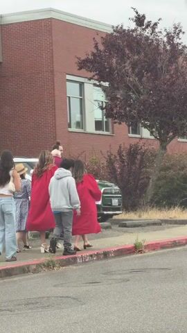 A group of people are walking along a sidewalk next to a brick building. The people are wearing red gowns and hats, suggesting they may be graduating. Some of the people are carrying objects, such as a skateboard and a purse. The environment appears to be an outdoor urban setting with a tree and a sidewalk.