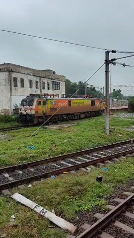 A train locomotive is parked on a track next to a building and a smaller structure. The locomotive is orange and green with "INDIAN RAILWAYS" written on its side. Overhead wires run across the top of the frame. The ground is covered in grass and gravel, with several sets of train tracks visible. The sky is overcast.